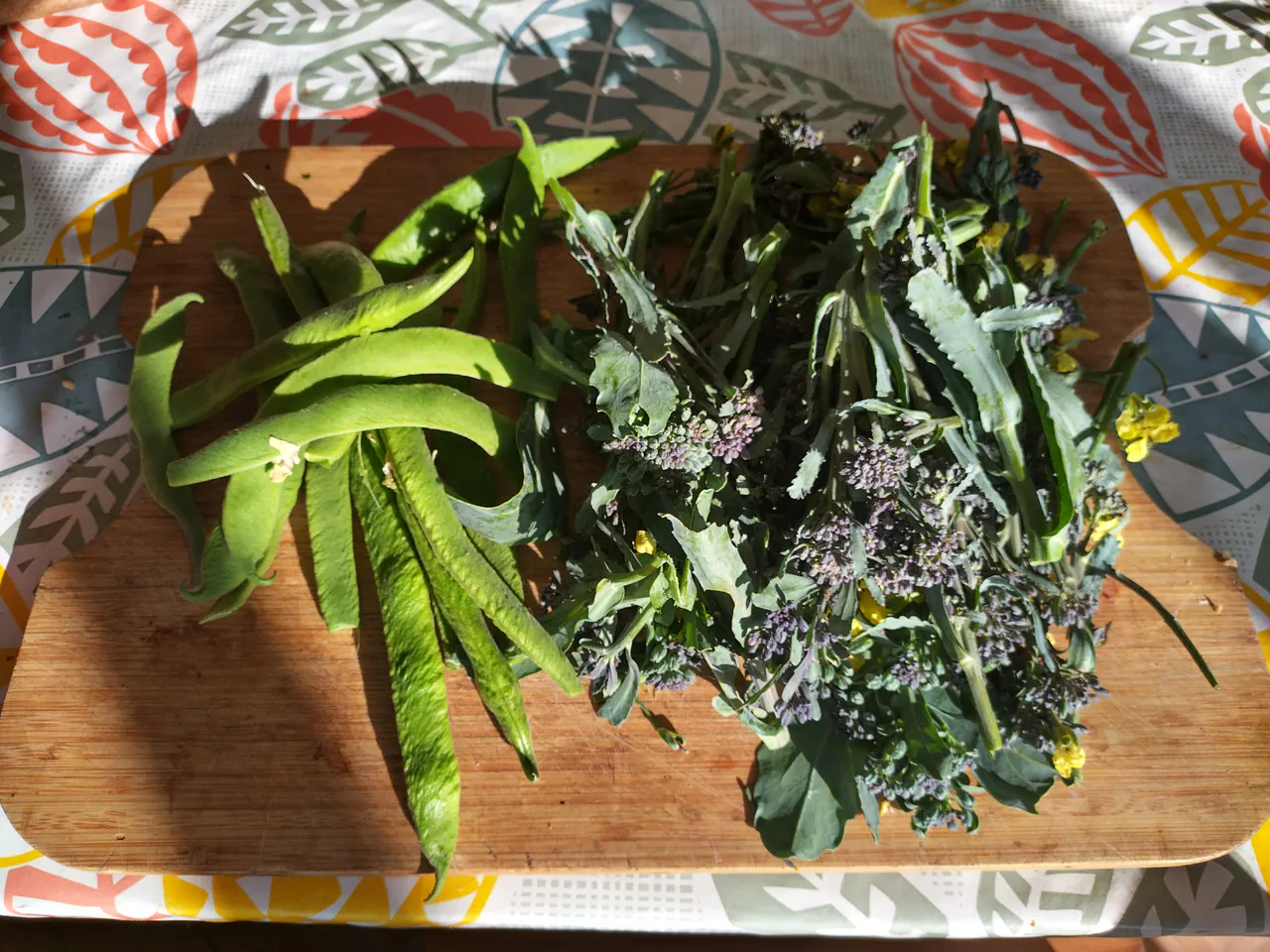 Fresh green beans and purple sprouting broccoli with yellow flowers arranged on a wooden chopping board